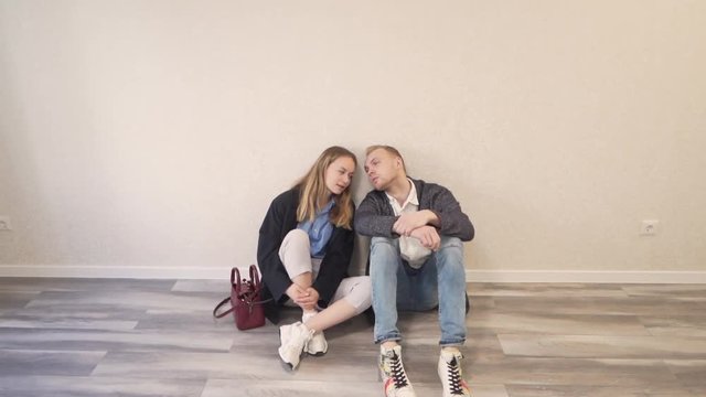Young Caucasian Married Couple Sitting on Floor In New Empty Finished Real Estate Property Leaning on Each Other and Talk. Light Camera Movement
