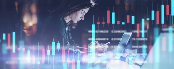 Business woman sitting at night office in front laptop computer and using her mobile phone. Red and green candlestick chart and stock trading computer screen background. Double exposure. Wide