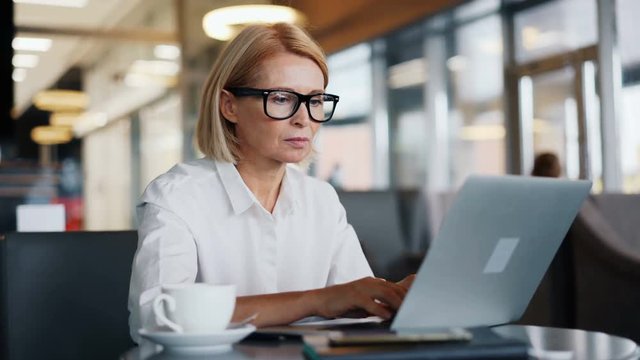 Busy entrepreneur senior woman wearing glasses is working with laptop typing in modern cafe concentrated on activity. Technology, career and people concept.