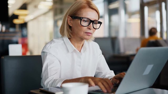 Corporate manager serious mature lady in white shirt is working with laptop in cafe typing business e-mail looking at computer screen. Career and technology concept.