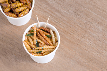 Bamboo Caterpillar and Silkworm Pupae insects for eating as food deep-fried snack in disposable cup for take out home on wood table background, is good source of protein edible. Entomophagy concept.