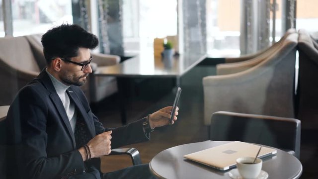 Entrepreneur serious man in glasses is making video call online using smartphone with camera in cafe sitting at table talking and gesturing. Conversation and Internet concept.
