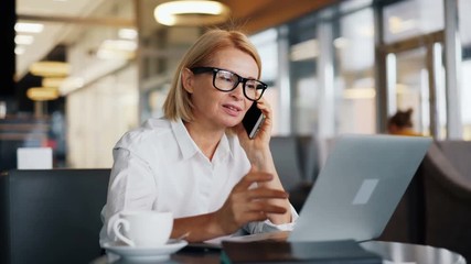Cheerful female entrepreneur smiling mature woman is speaking on mobile phone and using laptop working in cafe. Communication and business people concept. - Powered by Adobe