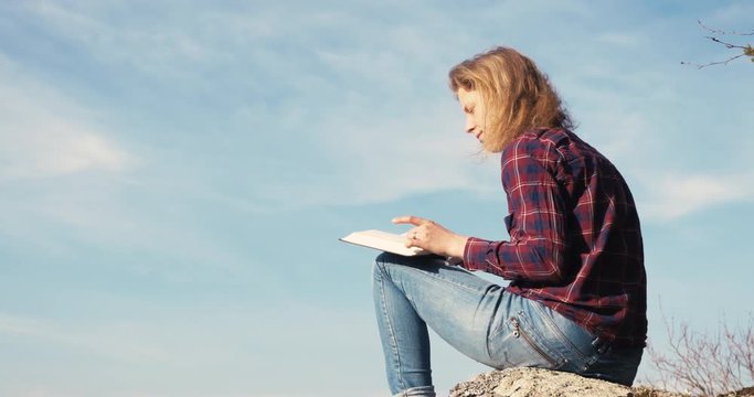 Girl reading and flipping pages in The Bible on a rock at a lake.