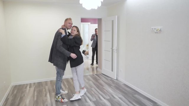 Young Caucasian Couple Celebrates Buying New Apartment Real Estate Property in Empty Room Spinning Around White Realtor Taking Pictures of Them With Smartphone