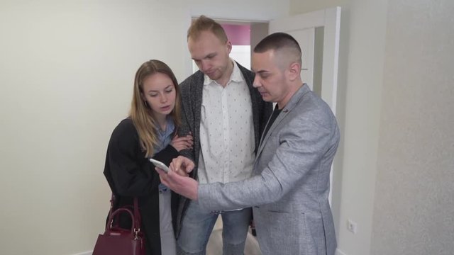 Young Caucasian Couple Standing in Emprty Room of New Apartment Property With Realtor Real Estate Broker Who Shows Them Content on Smartphone