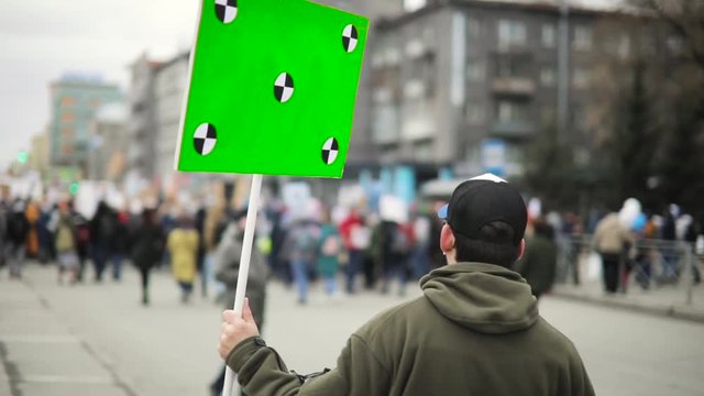 Revolution and demonstration in city during day. Man holds poster with green screen and walking the street with crowd on city rally, back view.