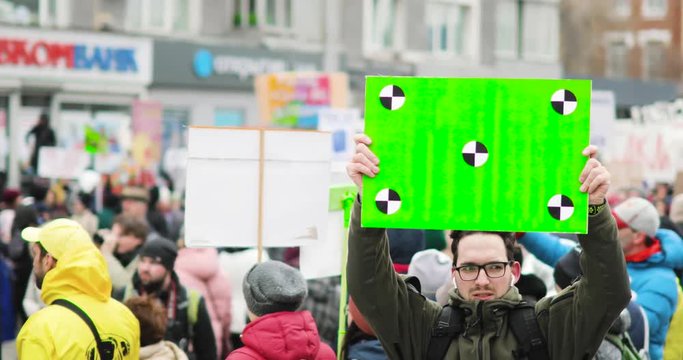 Young man stands and holds poster with green screen over his head on crowd background on the street during demonstration in the city, front view.