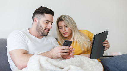 Young casual relaxed couple at home using smartphone and tablet sitting on a sofa. Man and woman with digital online connected devices. Generation Y relationship with technology concept.