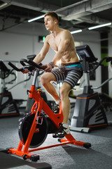 Fototapeta na wymiar Attractive man biking in the gym, exercising legs doing cardio workout cycling bikes