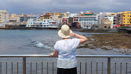 Middle aged Caucasian woman wearing a straw hat, looking towards the popular resort of El Medano, Tenerife, Canary Islands, Spain, concept for solo female adult traveling or travel vacation retirement