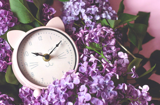 Pink alarm clock in the form of a cat lying on a bouquet of lilacs.