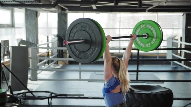 Three quarter back view tilt up of strong female athlete wearing weightlifting belt doing clean and jerk exercise in gym