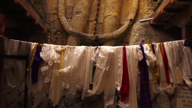 Extreme close up tilting shot of coloured khata scarfs hang by a string below a chamber statue. A tourist walks in to view the carved Buddha monument up on a rock surface, Mulbekh monastery, Ladakh