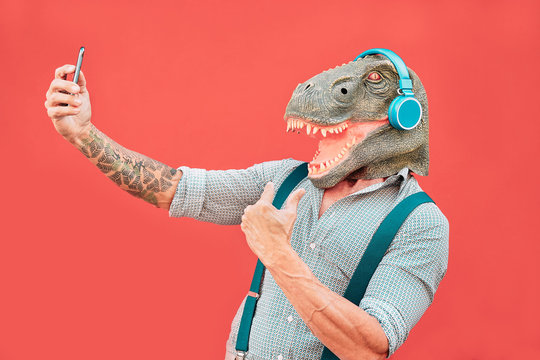 Crazy senior man wearing t-rex mask and taking selfie with mobile smartphone - Hipster older male having fun listening music and dancing outdoor -  Absurd, funny and surreal concepts