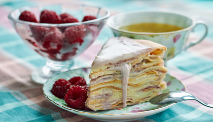Homemade layer cake with raspberry vanilla custard dripping decorated with frozen berry, table setting: cups of tea, spoons, dessert and ice-cream bowl with frozen fruit, close up, copy space