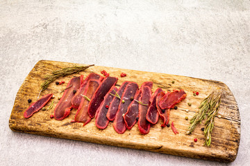 Basturma, dried tenderloin of beef meat, jerky, thinly sliced. Dry rosemary, pepper mix on vintage wooden board. Delicious food on a stone background.