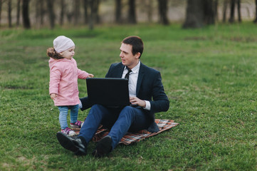 Fototapeta premium A serious businessman in a suit sits in a park on green grass and works on a laptop, a small child runs alongside and distracts. Computer, lifestyle, work. Father and daughter, business.