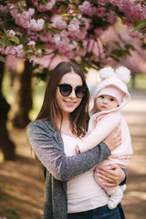 Fototapeta premium Beautiful mom with her little baby girl. Mom in sunglasses. Background of pink tree