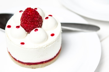 White airy cake with raspberry layer and raspberry berry lies on a white round plate next to a white cup, which stands on a white table