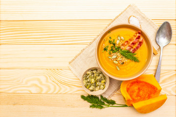 Homemade autumn hot pumpkin cream soup with smocked bacon and seeds. Raw pumpkin, fresh chives, dill, salt in spoon, vintage linen cloth on wooden background, top view, copy space.