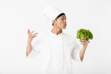 Image of asian young chief man in white cook uniform smiling at camera while holding green lettuce salad