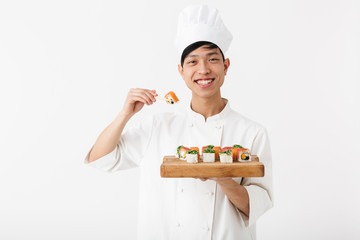 Image of positive chinese chief man in white cook uniform holding plate and eating sushi set with chopsticks