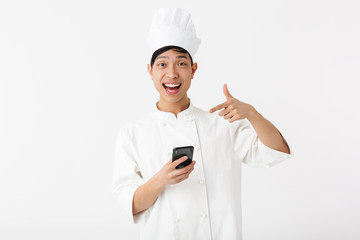 Image of young chinese chief man in white cook uniform and chef's hat holding mobile phone