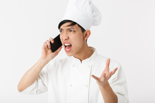Image of angry chinese chief man in white cook uniform and chef's hat talking on mobile phone