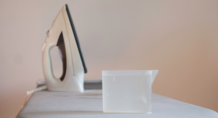 Plastic container with water and electric iron on ironing board 
