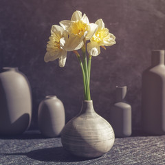 The flowers of narcissus are in a gray vase on a dark stone background. There are other vases of various sizes and shapes.