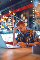 Handsome Caucasian blogger in plaid shirt and with serious face looking at agenda and holding hand on forehead. Cafe interior.