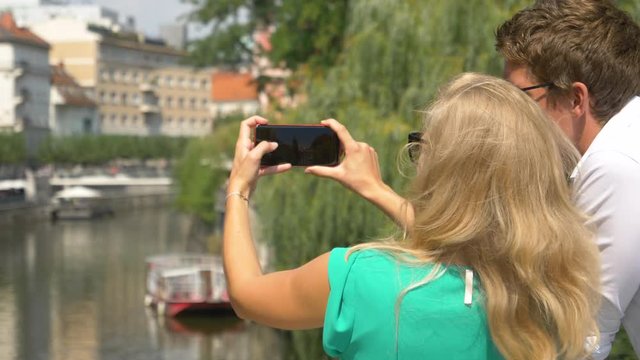 CLOSE UP SLOW MOTION DOF: Unrecognizable young blonde haired woman taking photos of the river with her new smartphone as she wanders around the scenic streets of Ljubljana with her cheerful boyfriend.