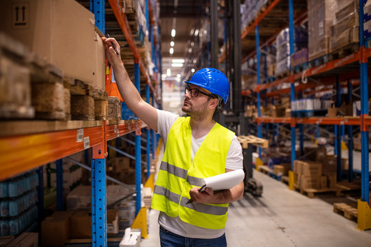 Professional warehouse worker in protective work wear holding checklist and checking inventory in storage room.