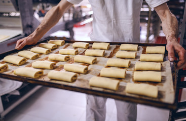 Caucasian baker in white uniform holding baking pray with raw sweet delicious pastry while standing in bakehouse.