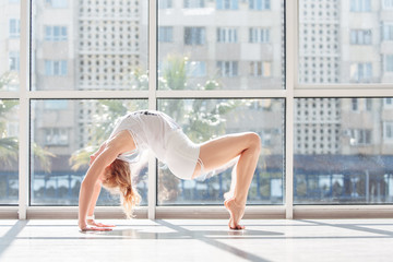 Slim beautiful young woman in a sports swimsuit doing chakrasana in the gym against the window. The concept of yoga at home