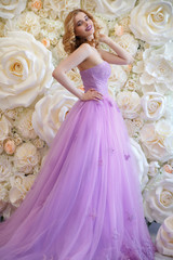 An elegant bride in a magnificent lilac dress stands near the photo zone of large white roses.