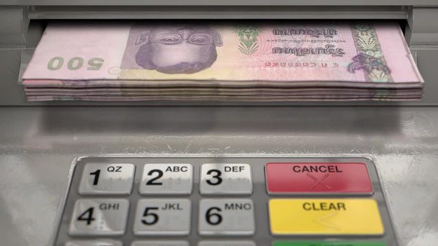 A closeup view of an atm facade with an illuminated sceen and keypad and a wad of thai bhat banknotes being ejected from the cash slot 