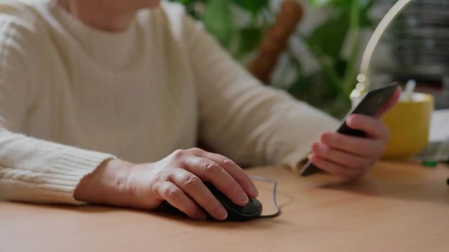 Senior woman working with gadgets at work space, focus on hand clicking on mouse, mobile phone in other hand. Grandmother using computer and smartphone. Old lady browsing internet, taking cellphone