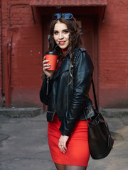 Portrait of a young happy curly brown-haired european girl dressed in red dress leather jacket holding coffee cup while standing in courtyard of old city. 