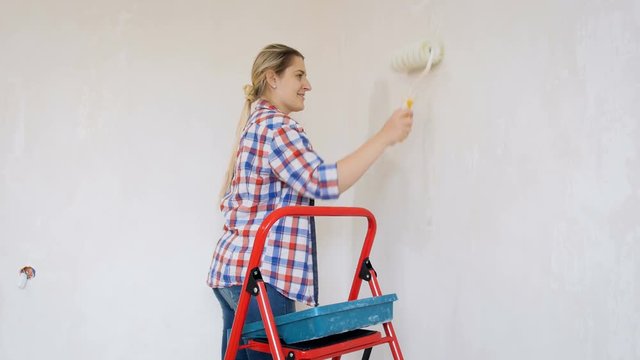 Video of beautiful young woman in checkerd shirt doing renovation at her house. Young girl painting walls with paint roller