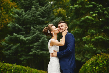 Beautiful young woman with perfect body and wedding hairstyle in lace dress hugs and kissing a handsome brutal man in the stylish blue suit outdoors at green park