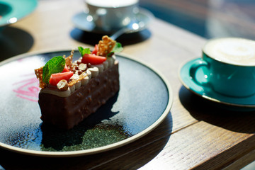 Cappuccino with frothy foam on wooden table near the window. Piece of chocolate cake with nuts and strawberries served with cup of coffee.