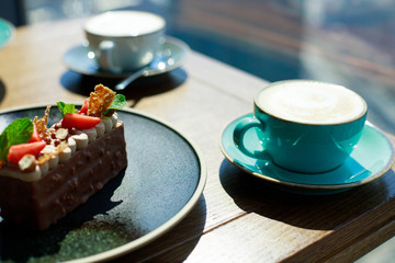 Cappuccino with frothy foam on wooden table near the window. Piece of chocolate cake with nuts and strawberries served with cup of coffee.