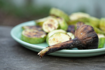 Grilled garlic head on a blue plate with grilled vegetable marrow on an old wooden table. Close up, soft focus