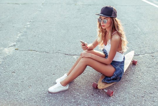 Beautiful girl in the summer in city, sitting on a skateboard, in the hands of the phone writes a message. Free space for text. The concept of online, application in social networks.