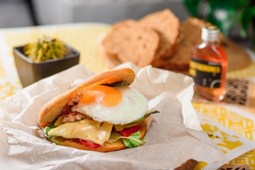 Burger with fried egg, chicken, cheese, tomatoes and other vegetables on a craft paper on yellow background in a restaurant. Series of photos for the menu.