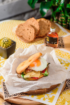 Burger with fried egg, chicken, cheese, tomatoes and other vegetables on a craft paper on yellow background in a restaurant. Series of photos for the menu.