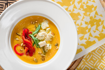 Pumpkin cream soup with cheese Dor Blue, bell pepper, pumpkin seeds in a white plate on yellow set on a wooden  table in a restaurant. close-up. top view. flat lay