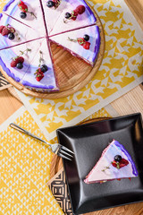 Beautiful layered berry cake with a purple, white and pink layer, decorated with raspberries and blueberries on top. Beautiful cake in a cut on a black saucer on a wooden table in the restaurant